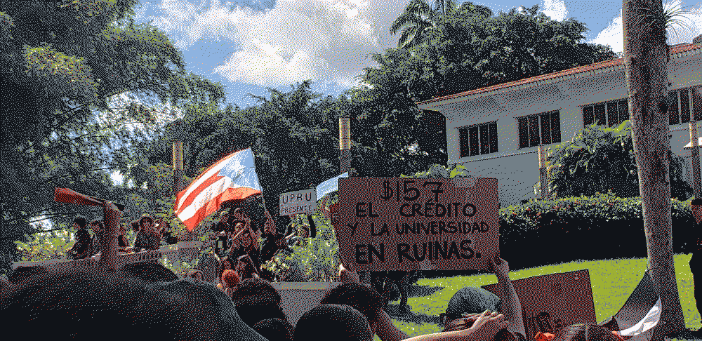 in front of the administration offices. the puertorican flag is seen in the background alongside a poster that reads '$157 el credito y la universidad en ruinas'