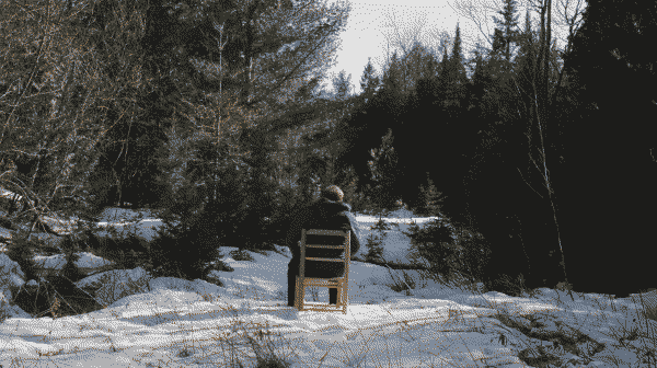 a screenshot from joe pera talks with you. its a shot of a snowy forest and in the center we see joe sitting, the camera is behind him.